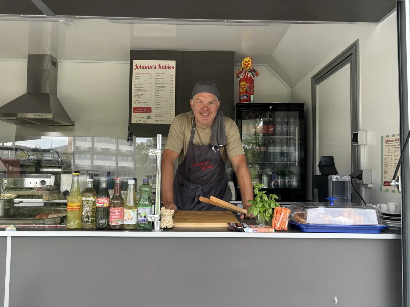 Ein Mann steht hinter einer Theke in einem Foodtruck, lächelt und trägt eine Schürze. Verschiedene Flaschen und ein Schneidebrett sind auf der Theke.