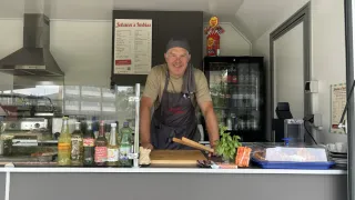 Ein Mann steht hinter einer Theke in einem Foodtruck, lächelt und trägt eine Schürze. Verschiedene Flaschen und ein Schneidebrett sind auf der Theke.