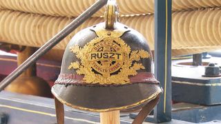Ein alter Militärhelm mit einem goldenen Emblem, das 'RUSZT' und 'POLIZEI' zeigt, auf einem Holzständer.
