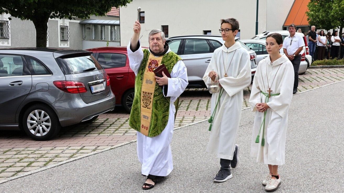 Ein Priester in grünen Gewändern und Sandalen führt eine Prozession mit zwei Akolythen in weißen Gewändern an, die ein Buch und einen silbernen Kelch halten, auf einem gepflasterten Bereich in der Nähe geparkter Autos.