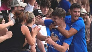 Eine Gruppe von Menschen in blauen Shirts feiert zusammen, wobei ein junger Mann in der Mitte lächelt und von anderen umarmt wird.