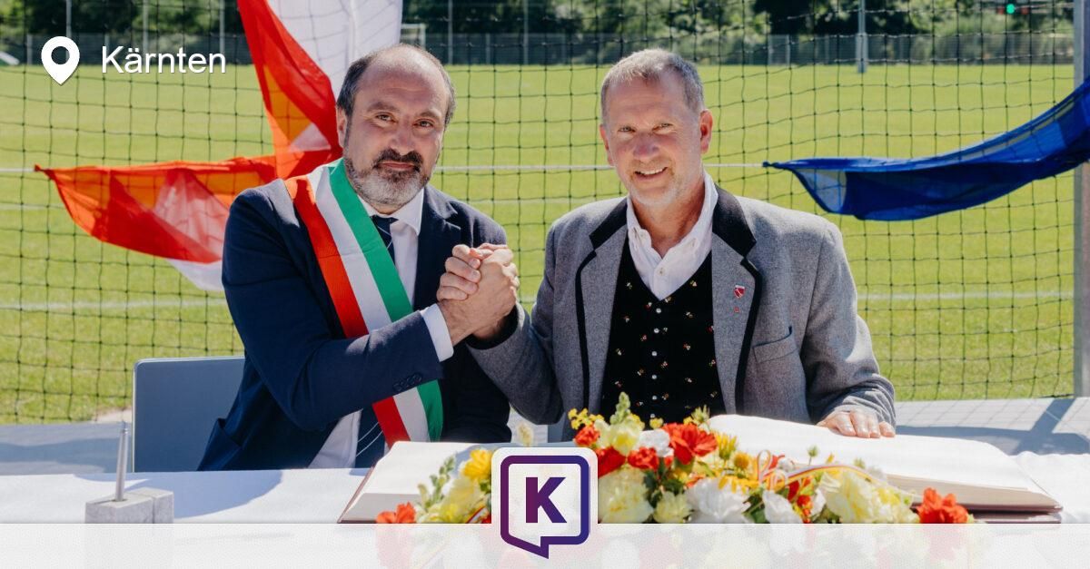 Zwei Männer sitzen an einem Tisch mit Blumen, einer trägt eine dreifarbige Schärpe, lächeln und schütteln Hände. Hinter ihnen ist eine Flagge und ein Netz zu sehen.