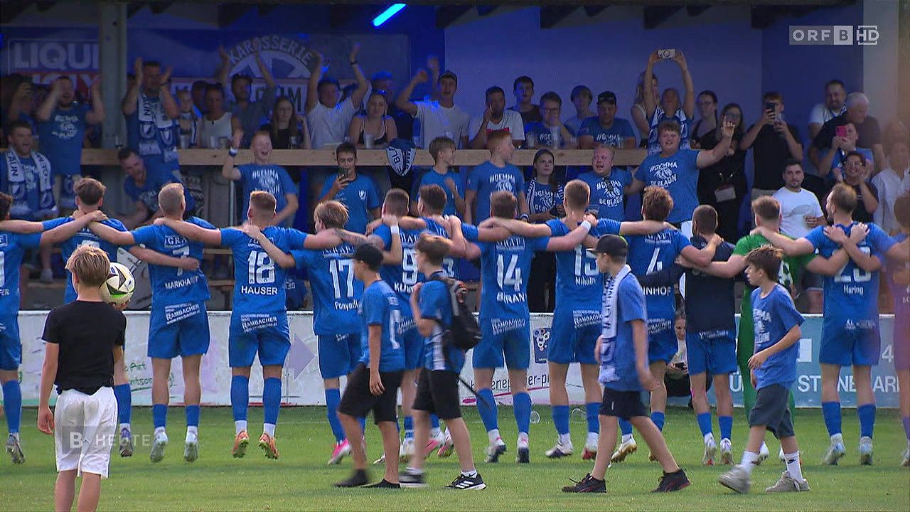 Eine Gruppe junger Athleten in blauen Trikots mit Nummern auf dem Rücken stehen auf einem Feld versammelt, möglicherweise um einen Sieg zu feiern. Dahinter jubelt eine Menge von Zuschauern, einige halten ihre Handys hoch.