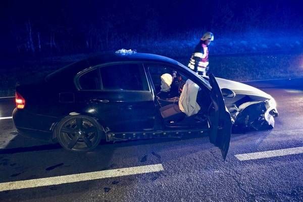 Ein schwarzer Wagen ist nachts an der Straßenseite beschädigt, mit einer Person im Inneren und einem Feuerwehrmann in der Nähe.