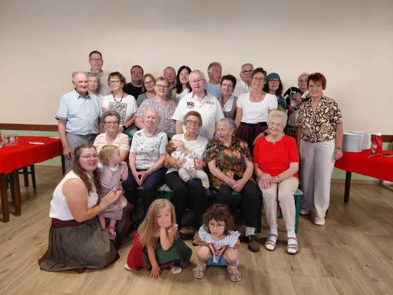 Eine Gruppe von Menschen, einschließlich Erwachsener und Kindern, posiert für ein Foto in einem Raum mit Holzboden. Einige sitzen, andere stehen. Ein Baby wird von einem Erwachsenen gehalten.