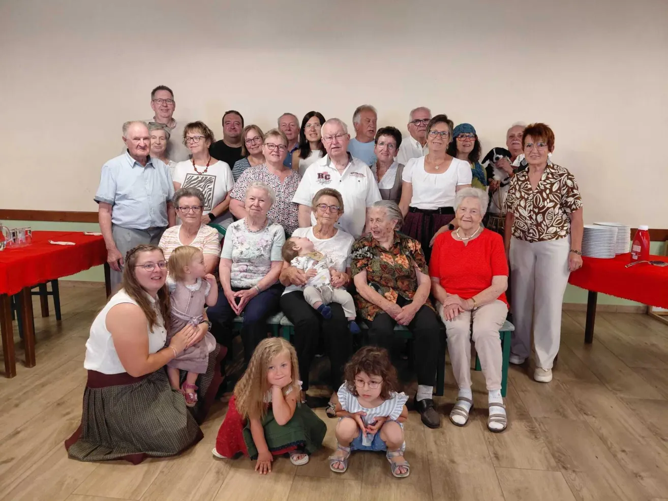 Eine Gruppe von Menschen, einschließlich Erwachsener und Kindern, posiert für ein Foto in einem Raum mit Holzboden. Einige sitzen, andere stehen. Ein Baby wird von einem Erwachsenen gehalten.