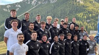 Eine Gruppe junger Männer in schwarzen und weißen Sportjacken mit Logo posiert für ein Foto vor einem Berg.