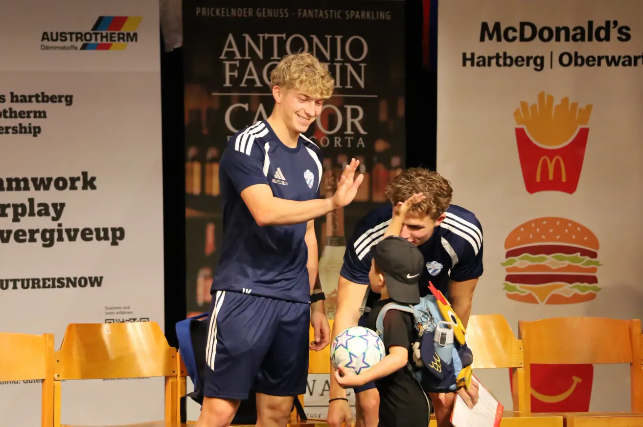 Zwei junge Fußballspieler in blauen Trikots interagieren mit einem Kind, das einen Fußball hält. Sie stehen vor einem McDonald's-Werbebanner.