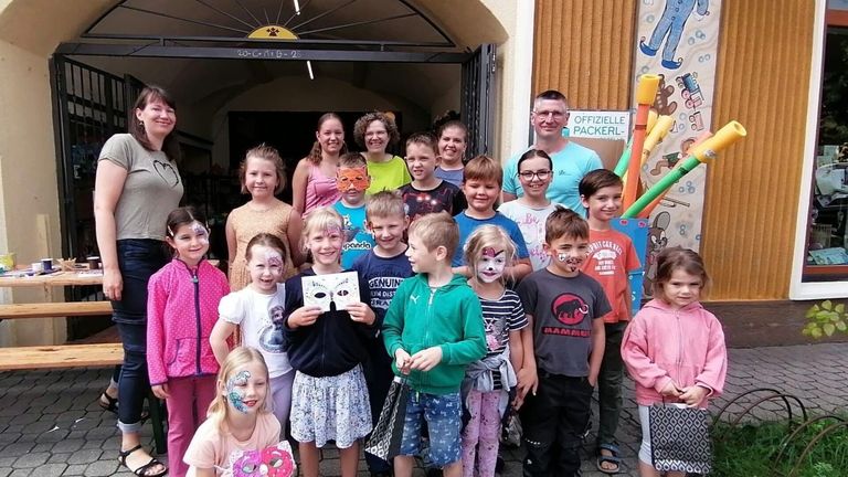 Eine Gruppe von Kindern und Erwachsenen mit Gesichtsbemalung, die vor einem Gebäude mit einem Schild stehen, auf dem 'Offizielle Packerl' steht. Die Kinder lächeln und posieren für ein Foto.
