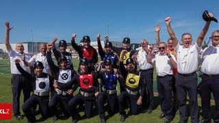 Eine Gruppe von Feuerwehrleuten in Uniformen und Helmen posiert für ein Foto auf einem Feld, wobei einige ihre Hände heben und andere lächeln. Im Hintergrund ist eine verschwommene Ansicht eines Stadions und einer Straßenlaterne zu sehen.