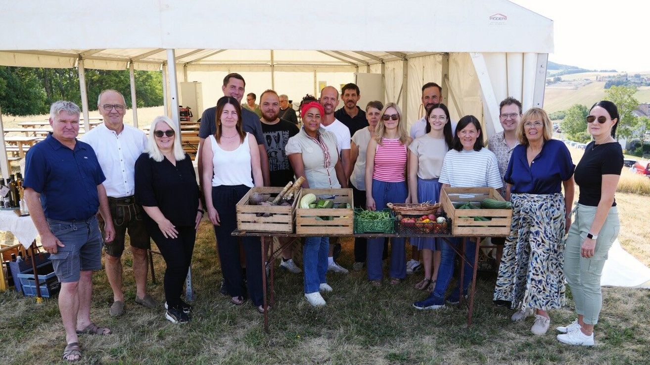 Eine Gruppe von Menschen steht unter einem Zelt und lächelt für ein Foto. Sie sind von Holzkisten voller Gemüse umgeben. Die Umgebung scheint ein Markt oder eine Outdoor-Veranstaltung zu sein.