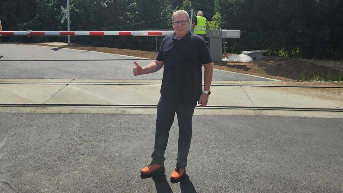 Ein Mann in einem schwarzen Shirt und einer Brille steht auf einer Straße und gibt einen Daumen hoch. Hinter ihm ist eine Schranke mit einem roten und weißen Streifen.