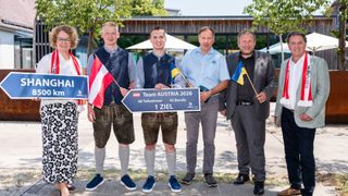 Fünf Männer stehen auf einem Bürgersteig, zwei halten Flaggen und drei halten ein Schild mit der Aufschrift 'Team Austria 2026'. Sie sind in traditioneller Kleidung und Geschäftsanzügen gekleidet.