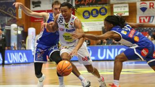 Drei Basketballspieler in Aktion auf einem Hallenplatz. Der Spieler in der Mitte dribbelt den Ball, während die anderen versuchen, ihn zu blockieren.