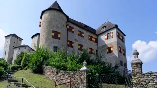Ein großes mittelalterliches Schloss mit roten und gelben Fensterläden und einer Steinmauer davor.