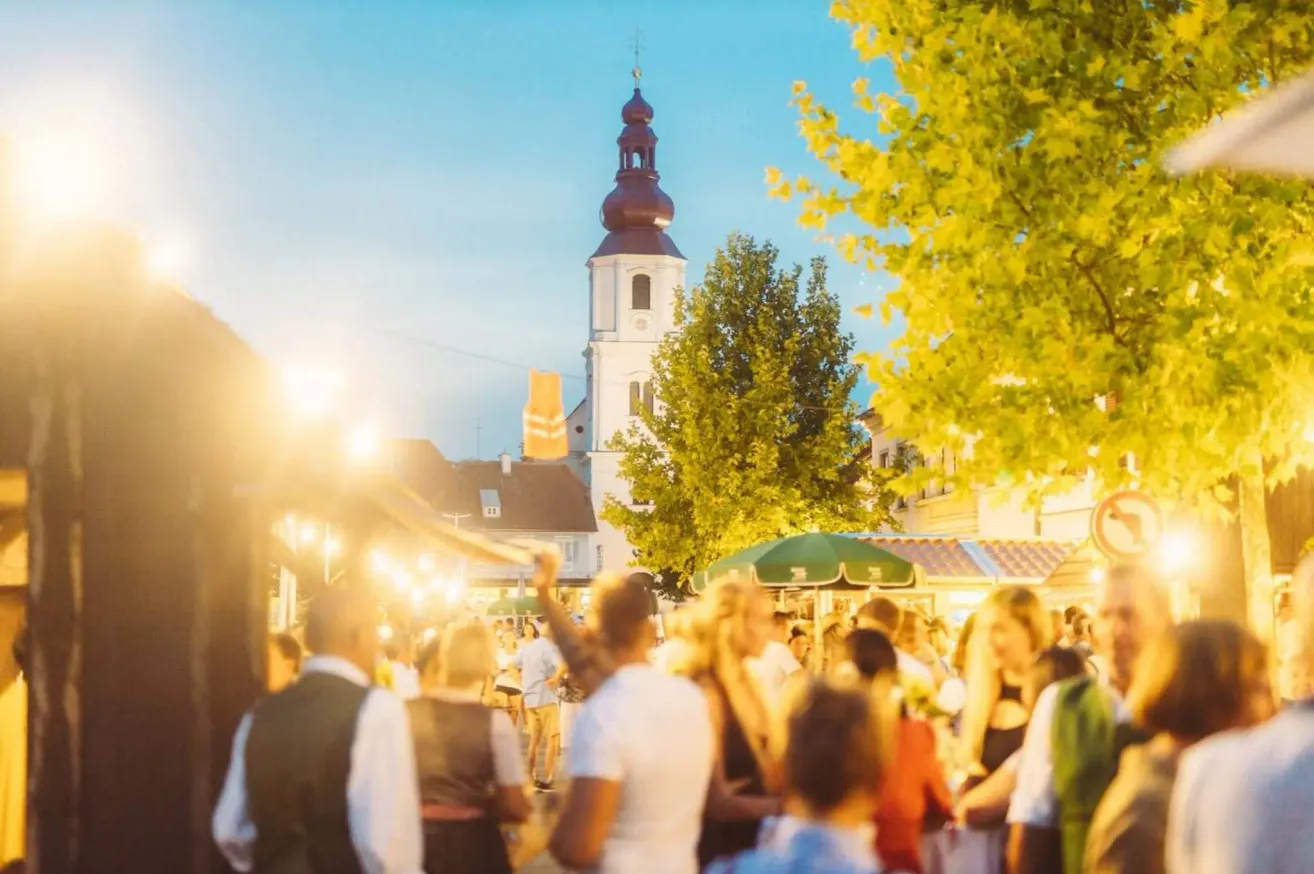 Ein geschäftiger Outdoor-Markt bei Dämmerung mit Menschen, die sich mischen, einem Kirchturm im Hintergrund und einem grünen Schirm, der Schatten spendet.