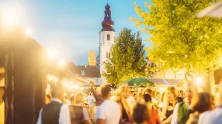 Ein geschäftiger Outdoor-Markt bei Dämmerung mit Menschen, die sich mischen, einem Kirchturm im Hintergrund und einem grünen Schirm, der Schatten spendet.