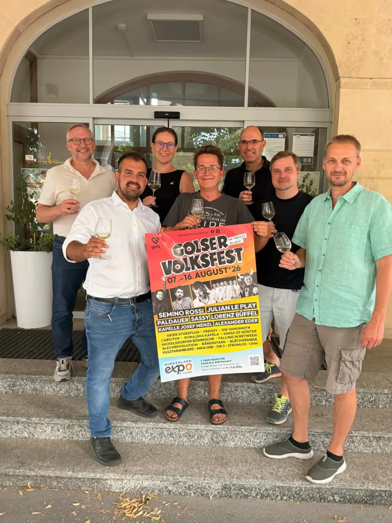 Eine Gruppe von Menschen steht vor einem Gebäude, hält Weingläser in der Hand und lächelt. Sie halten ein Plakat mit dem Text 'Golser Volksfest' und einem Datumsbereich. Der Mann in der Mitte hält ein Plakat mit dem Text 'Golser Volksfest' und einem Datumsbereich.