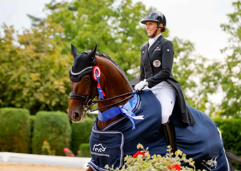 Eine Frau in Reiterausrüstung reitet ein braunes Pferd mit einem blauen Cape, trägt einen Helm und Handschuhe, mit einem blauen Band am Pferdekopf.