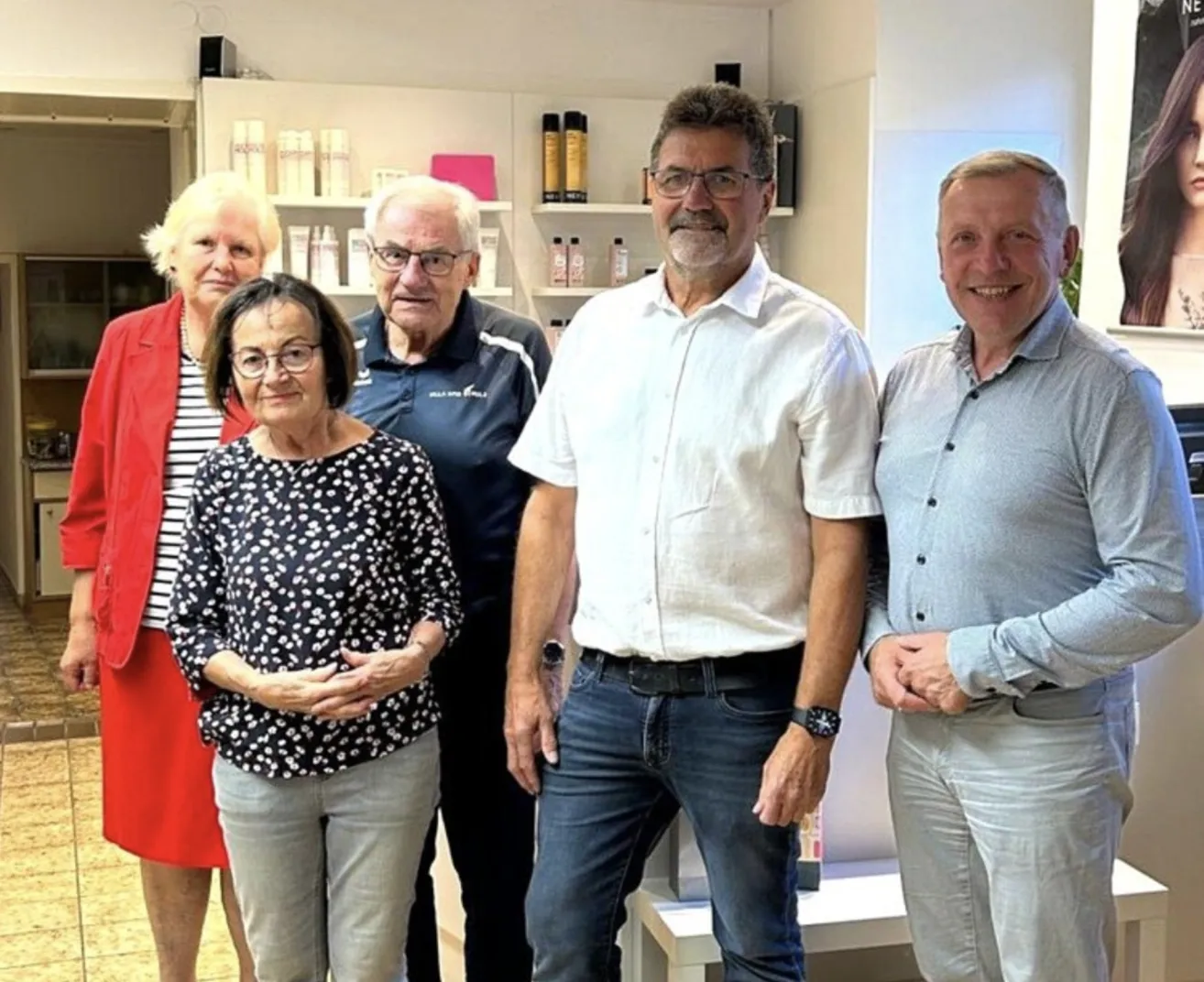 Fünf Personen stehen in einem Raum und lächeln für ein Foto. Drei Männer und zwei Frauen. Der Mann in der Mitte trägt ein weißes Hemd und Jeans. Der Mann rechts trägt ein blaues Hemd und Jeans. Die Frau links trägt eine rote Jacke und eine Halskette.