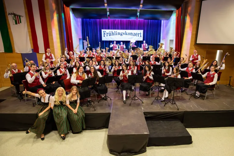 Ein großes Orchester auf der Bühne, mit Musikern, die verschiedene Instrumente spielen, und drei Frauen in traditionellen Kostümen davor sitzend. Die Bühne ist mit einem Banner mit der Aufschrift 'Frühlingskonzert' dekoriert.