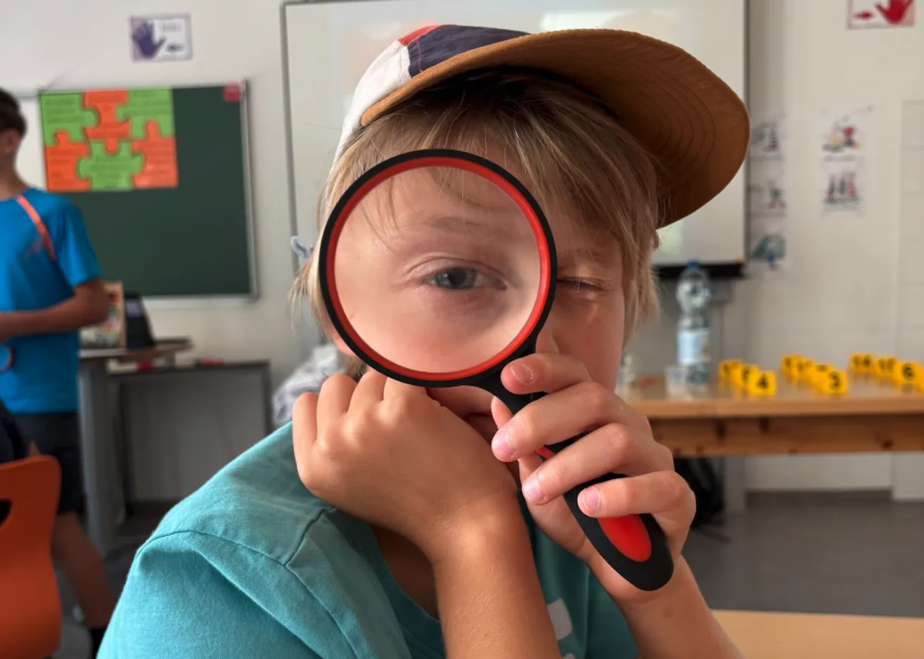 Ein junger Junge mit Hut und blauem Shirt hält eine Lupe dicht vor sein Gesicht und schaut aufmerksam hinein.
