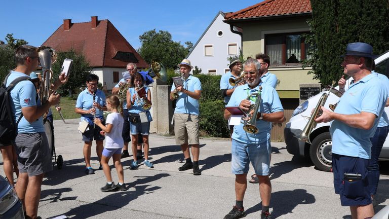 Ein Blasorchester spielt auf einer Dorfstraße, mit Musikern, die Trompeten, Posaunen und andere Instrumente spielen. Ein junges Mädchen geht vor ihnen.