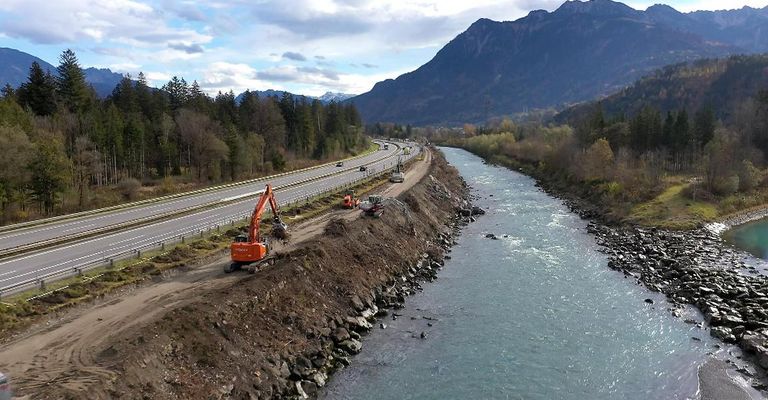 Eine Autobahn mit fahrenden Autos neben einem Fluss mit Felsen und einer Bergkette im Hintergrund. Baumaschinen sind am Straßenrand vorhanden.