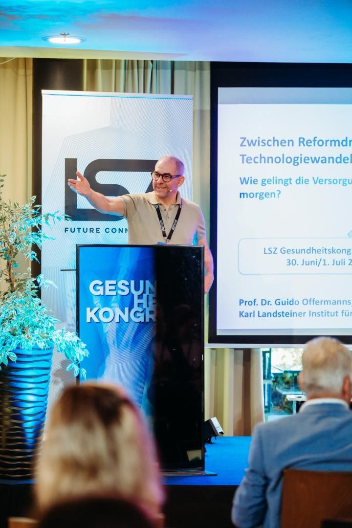 Ein Mann spricht an einem Podium mit einem blauen Hintergrund, der mit 'Gesundheitskongress' beschriftet ist. Ein Bildschirm hinter ihm zeigt eine Präsentation mit dem Titel 'Zwischen Reformdr Technologiewandel'. Eine Pflanze in einem blauen Topf ist links.