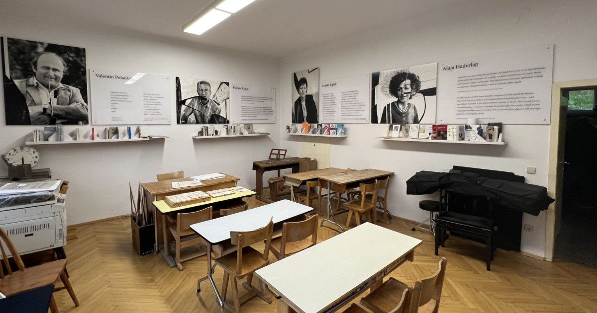 Ein Klassenzimmer mit Holztischen und Stühlen, einem Klavier und einer Wand mit gerahmten Bildern und Text.