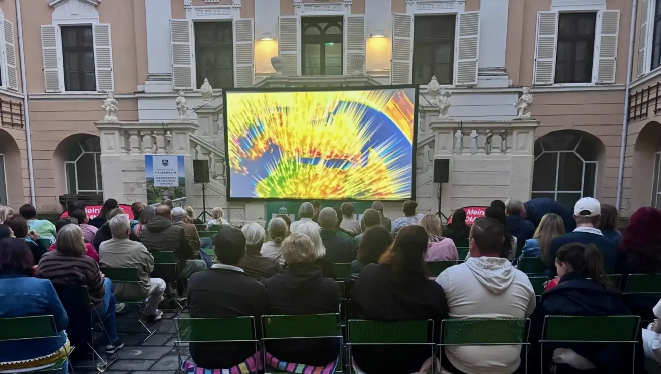 Eine Menschenmenge schaut auf eine große Leinwand, die ein farbenfrohes Bild vor einem Gebäude zeigt.