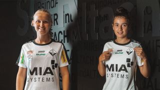 Zwei Frauen in weißen Fußballtrikots mit Logos und Texten stehen nebeneinander vor einer Wand mit Text.
