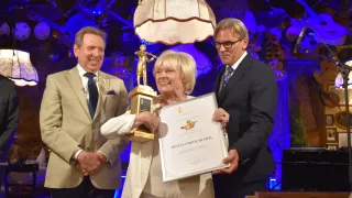 Eine Frau in einem weißen Anzug hält eine goldene Trophäe und ein Zertifikat in der Hand, während sie zwischen zwei Männern in formeller Kleidung steht, alle lächeln und für ein Foto posieren.