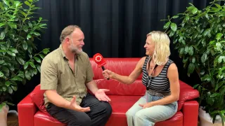 Ein Mann und eine Frau sitzen auf einem roten Sofa, die Frau hält ein Mikrofon und interviewt den Mann.