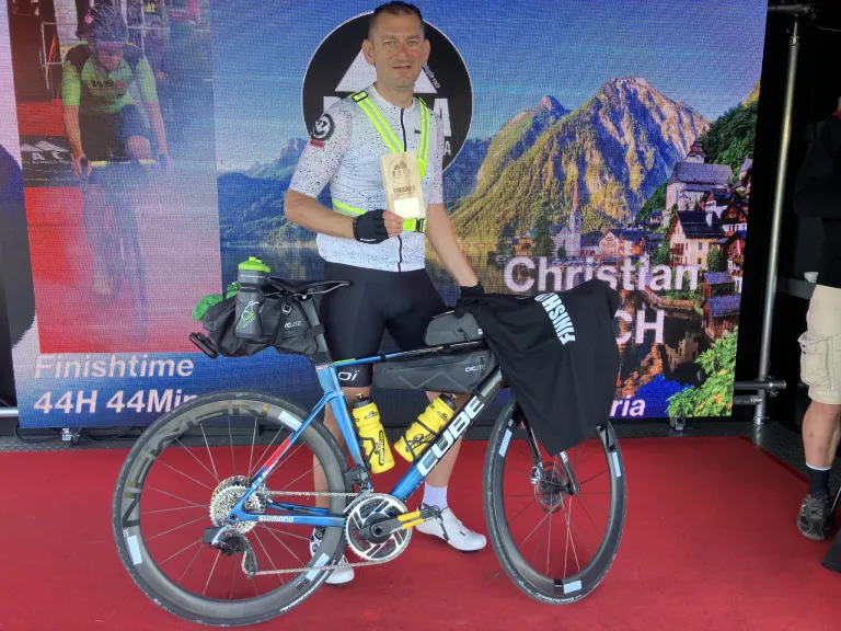 Ein Mann steht auf einem roten Teppich mit seinem Fahrrad, hält eine Medaille und trägt eine reflektierende Weste. Das Fahrrad hat Wasserflaschen und eine Tasche. Dahinter ist eine große Leinwand mit einer Berglandschaft.