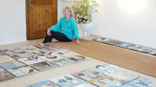 Eine ältere Frau sitzt auf einem Teppich vor einer Wand, lächelt und ist von fertigen Gemälden von Frauen umgeben.