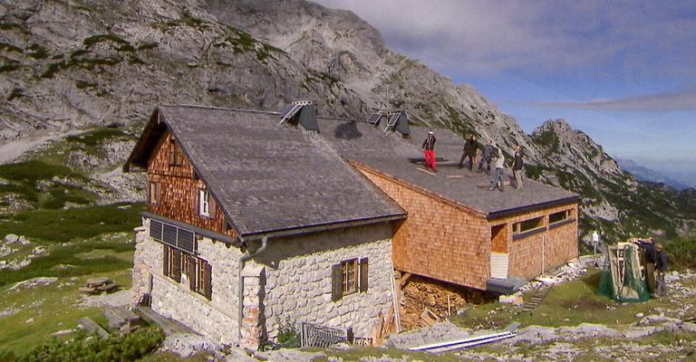 Eine Gruppe von Menschen arbeitet an einem Steinhaus mit grauem Dach und Solarpanelen, das sich auf einem Berg mit bewölktem Himmel befindet.