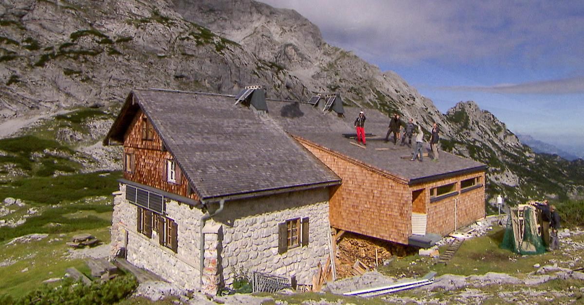 Eine Gruppe von Menschen arbeitet an einem Steinhaus mit grauem Dach und Solarpanelen, das sich auf einem Berg mit bewölktem Himmel befindet.