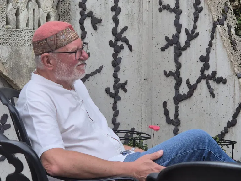 Ein älterer Mann mit weißem Bart und Brille sitzt auf einem Stuhl und trägt eine rosa Kopfband. Er lächelt und schaut nach vorne. Hinter ihm befindet sich eine Wand mit einem geometrischen Muster aus schwarzen Steinen.
