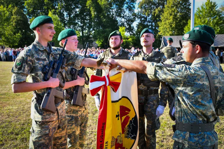 Fünf Soldaten in Militäruniformen halten eine Flagge und schütteln sich die Hände vor einer Menge.
