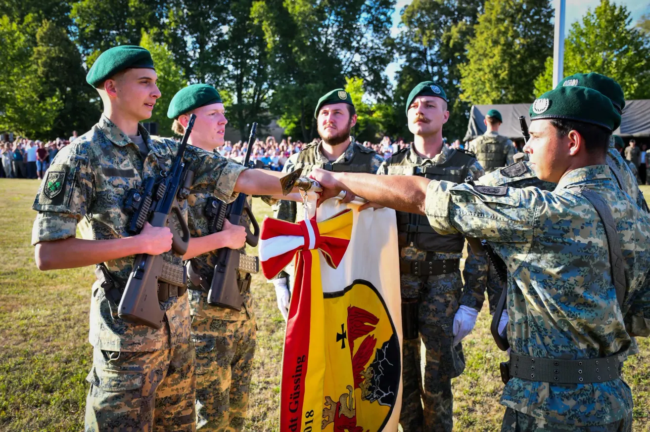 Fünf Soldaten in Militäruniformen halten eine Flagge und schütteln sich die Hände vor einer Menge.