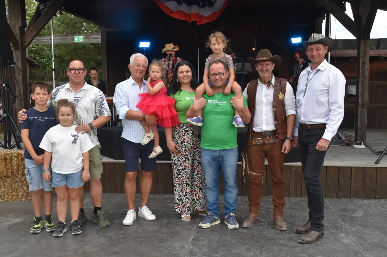 Eine Gruppe von Menschen, einschließlich Erwachsener und Kindern, posiert für ein Foto auf einer Bühne mit einem Hintergrund einer amerikanischen Flagge und einer Band, die im Hintergrund spielt.