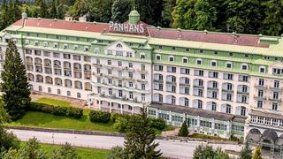 Ein Luftbild eines großen Hotels namens Panhans, umgeben von üppigem Grün und Bäumen. Das Gebäude hat ein rotes Dach und grüne Akzente, mehrere Stockwerke und zahlreiche Fenster.