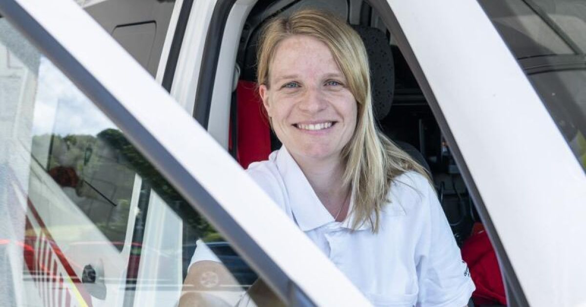 Eine Frau mit blonden Haaren sitzt in einem Fahrzeug, lächelt und trägt ein weißes Hemd. Sie ist wahrscheinlich eine Sanitäterin oder Notfallhelferin.