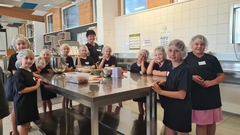 Eine Gruppe von Kindern mit Haarnetzen bereitet in einer Küche mit einer Frau hinter ihnen Essen zu.