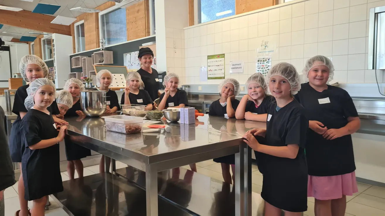 Eine Gruppe von Kindern mit Haarnetzen bereitet in einer Küche mit einer Frau hinter ihnen Essen zu.