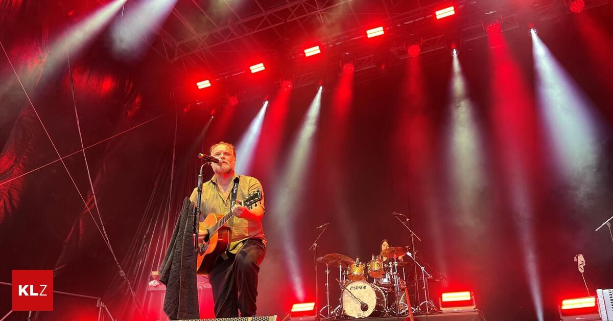 Ein Mann spielt Gitarre und singt auf der Bühne mit einem Schlagzeug und Becken hinter ihm. Die Bühne ist mit roten und weißen Lichtern beleuchtet.