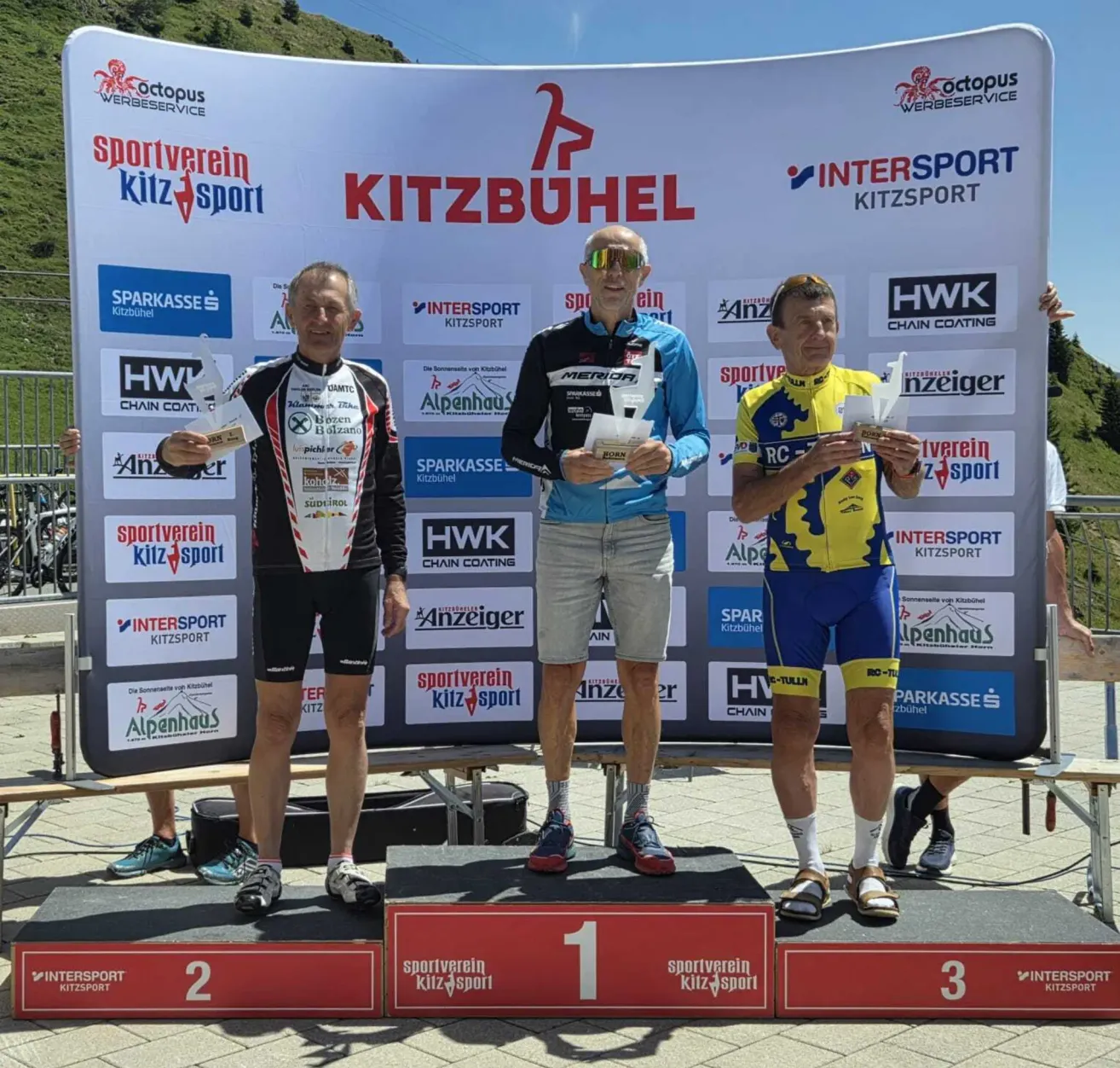 Drei Männer stehen auf einem Podium mit Medaillen, einer hält ein Papier, ein anderer hält eine Trophäe und der dritte hält ein Papier. Dahinter ist ein Banner mit Logos und Text.