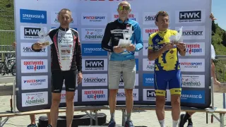 Drei Männer stehen auf einem Podium mit Medaillen, einer hält ein Papier, ein anderer hält eine Trophäe und der dritte hält ein Papier. Dahinter ist ein Banner mit Logos und Text.