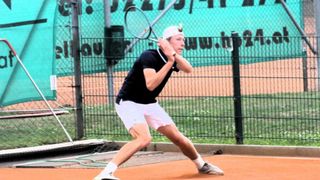 Ein Mann in einem schwarzen Shirt und weißen Shorts schwingt einen Tennisschläger auf einem Tennisplatz mit einem grünen Zaun und einer grünen Wand im Hintergrund.